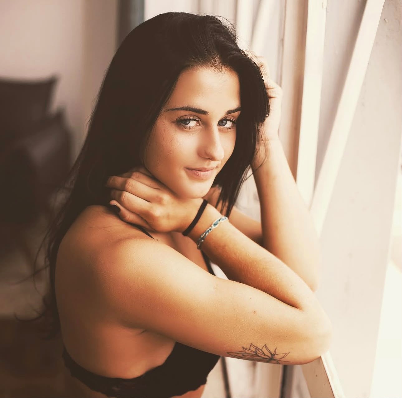 Retrato de uma mulher jovem de cabelos escuros e longos, apoiando-se perto de uma janela com iluminação suave e olhando para a câmera com um sorriso discreto.