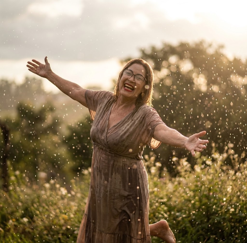 Mulher sorridente com cabelos castanhos e óculos de grau dançando ao ar livre sob uma chuva fina e iluminada pelo sol. Ela está de braços abertos em um gesto de liberdade, vestindo um vestido fluido, com um fundo verde natural desfocado e gotas de chuva brilhando como cristais no ar.