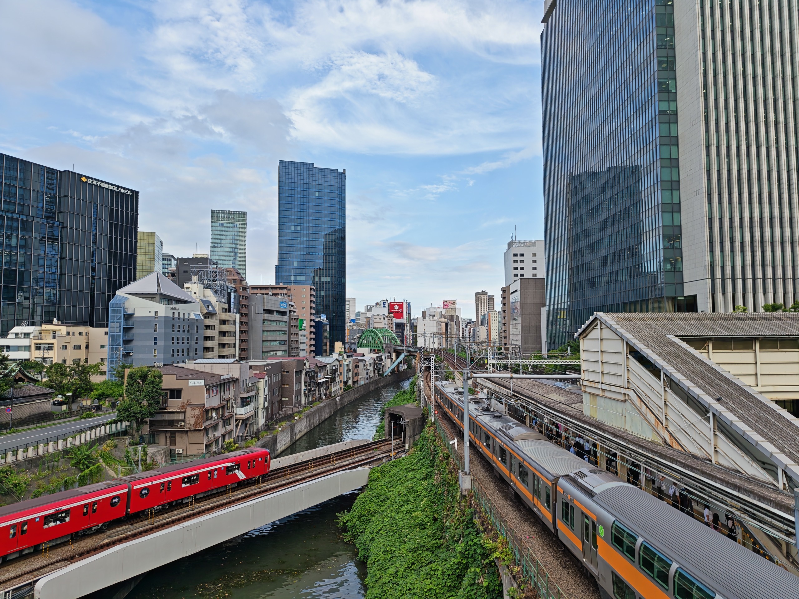 Fotografia diurna tirada da Ponte Hijiri em Ochanomizu, Tóquio, mostrando o rio Kanda cortando o cenário urbano. À direita, um trem laranja e cinza da linha Chuo na estação; abaixo, um trem vermelho do metrô linha Marunouchi cruza uma ponte sobre as águas, cercados por modernos edifícios espelhados sob um céu com nuvens.