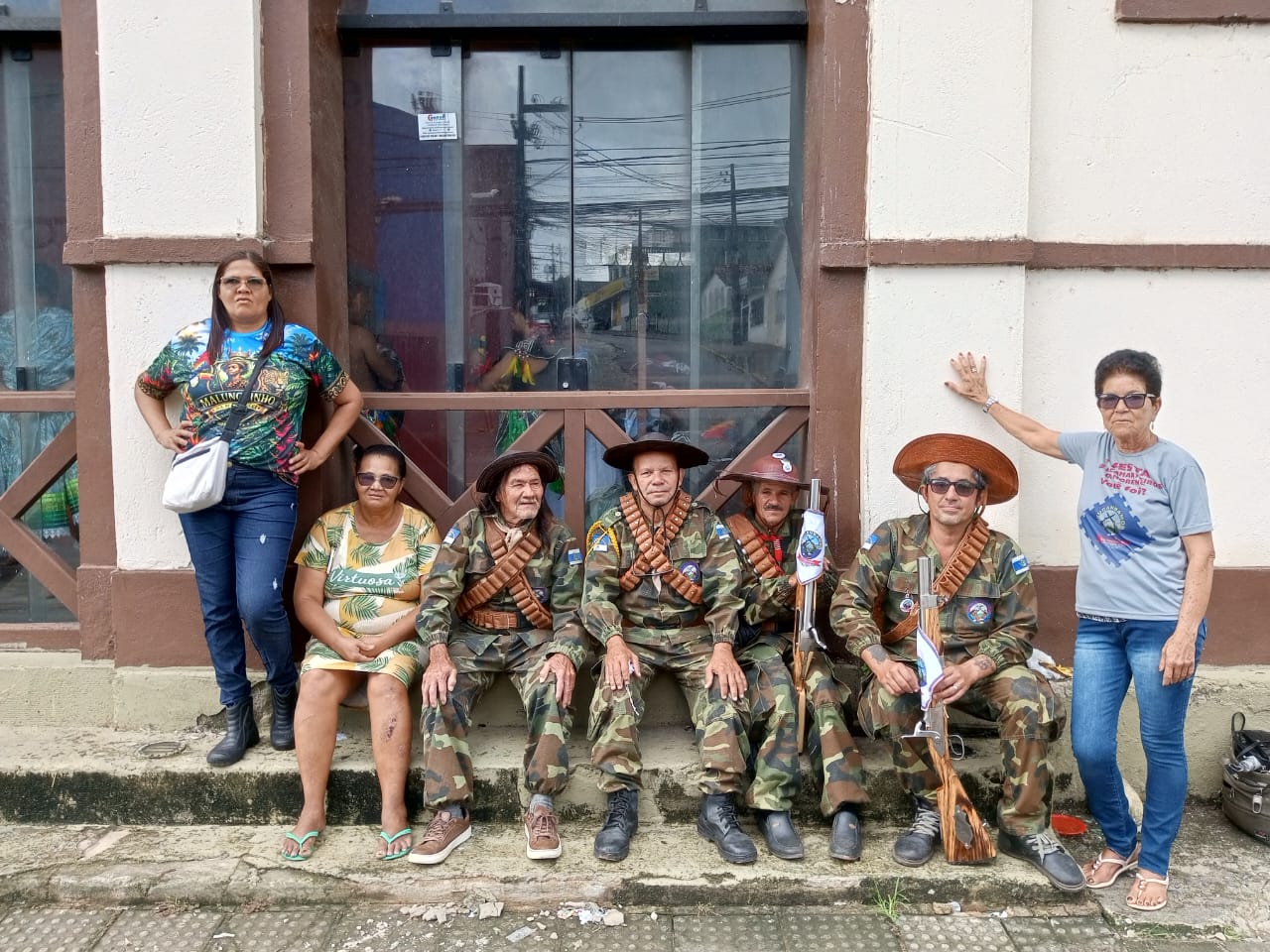 Fotografia em ambiente externo e luz do dia. Sete pessoas posam em grupo diante da calçada de um edifício de paredes claras com detalhes em pintura marrom e portas de vidro reflexivo. À esquerda: Uma mulher está em pé, vestindo calça jeans escuro, botas pretas e uma camiseta estampada colorida com a palavra "Malunguinho". Ao lado dela, uma mulher sentada veste um vestido amarelo estampado com folhas e a palavra "Virtuosa". Ao centro: Quatro homens estão dispostos lado a lado, três deles sentados e um levemente inclinado. Todos vestem fardamento camuflado no estilo militar, chapéus tradicionais de couro com abas dobradas e canotadeiras (cintos de munição de couro) cruzadas sobre o peito. Dois deles seguram réplicas de espingardas de madeira ou ornamentais. À direita: Uma mulher em pé apoia uma das mãos na parede. Ela usa óculos escuros, calça jeans azul, sandálias e uma camiseta cinza com estampa de evento.
