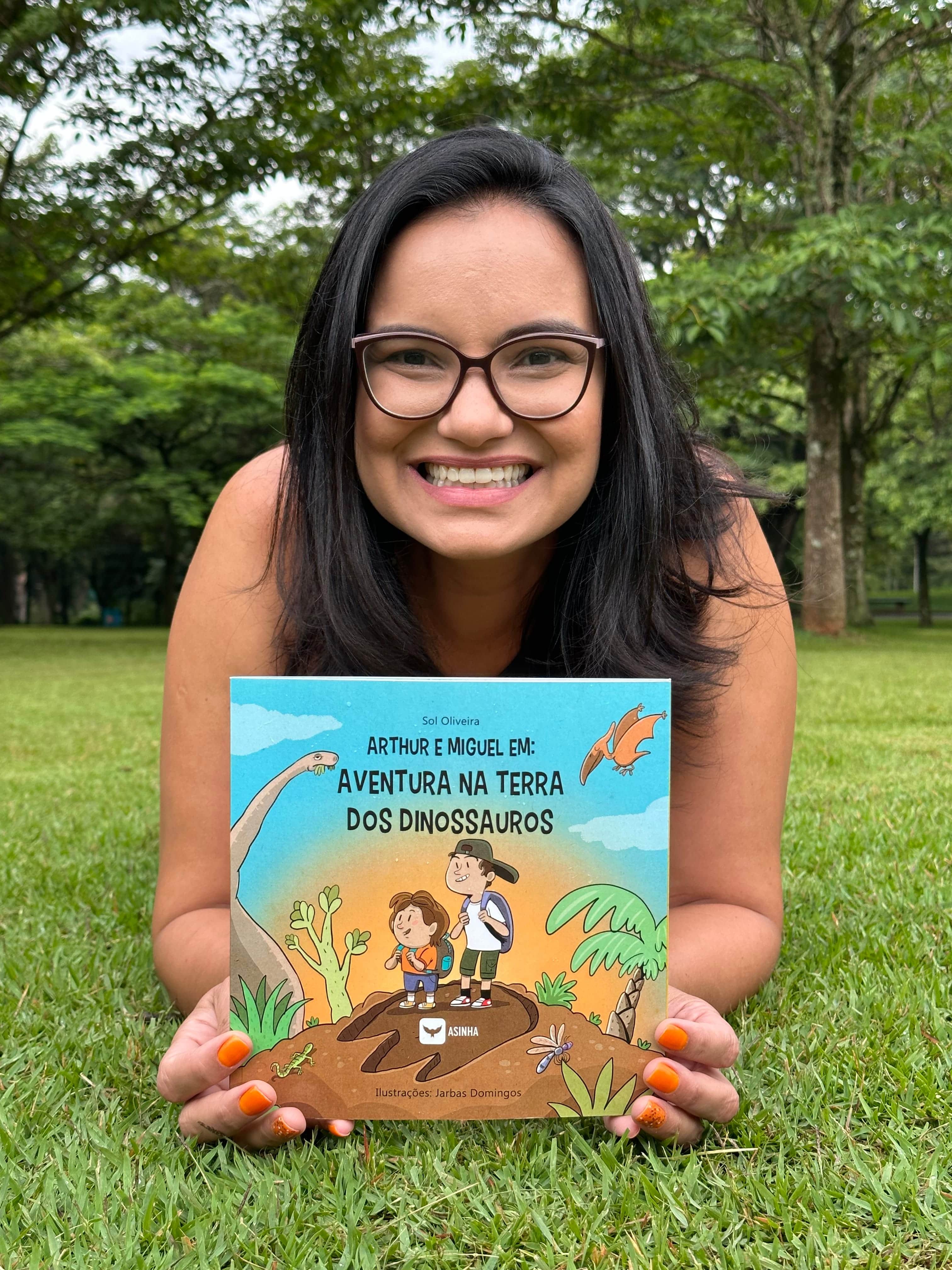 A autora Sol Oliveira, sorrido, deitada  na grama segurando o seu livro