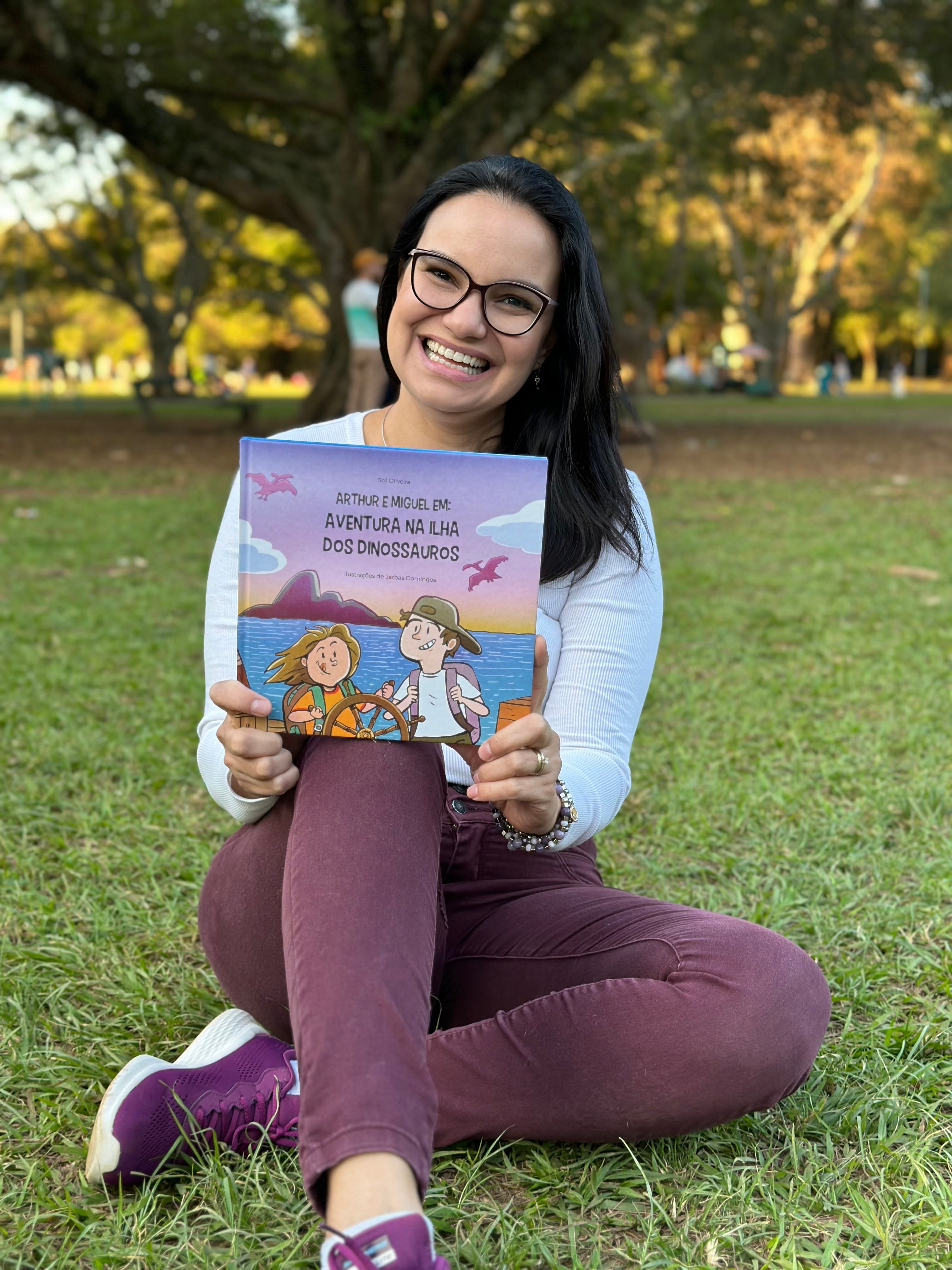 A autora Sol Oliveira, sorrido, sentada na grama com o livro na mão