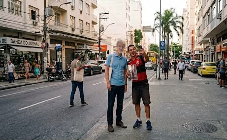Fotografia em plano médio e ao ar livre mostra o jornalista e escritor David Mitchell ao lado de um fã em uma rua movimentada.À esquerda, David Mitchell, um homem branco de cabelos claros e óculos, veste uma camisa polo azul-clara, calça escura e carrega uma ecobag no ombro. À direita, o fã, um homem jovem e sorridente, veste a camisa do time de futebol Flamengo e bermuda escura. Ele segura um livro com a mão direita e eleva o celular com a esquerda para tirar uma selfie dos dois. A cena se passa na calçada de uma avenida urbana com prédios residenciais e comerciais ao fundo, além de carros em movimento e pedestres circulando. Do lado esquerdo, é possível ver parte de uma banca de jornal.