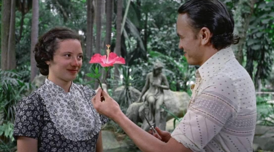 Uma cena de um filme de época em um jardim tropical. Um homem de camisa de botões estampada segura uma flor de hibisco rosa e a oferece a uma jovem com um sorriso tímido. Ela está usando um vestido escuro com uma gola de babados floral. Ao fundo, uma estátua clássica de pedra está sentada em uma lagoa de jardim.