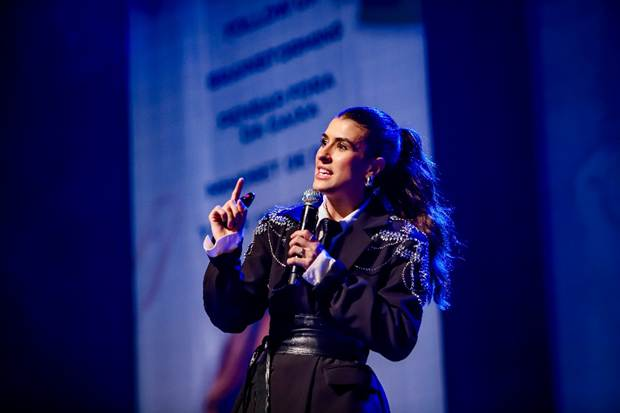 A humorista Luana Zucoloto no palco do Teatro AXIA Casa Grande, segurando um microfone e apontando o indicador para cima enquanto apresenta o espetáculo 'Esse Show Poderia Ter Sido um E-mail'. Ela veste um blazer preto com bordados brilhantes nos ombros, camisa social e gravata preta, sob iluminação cênica azul.