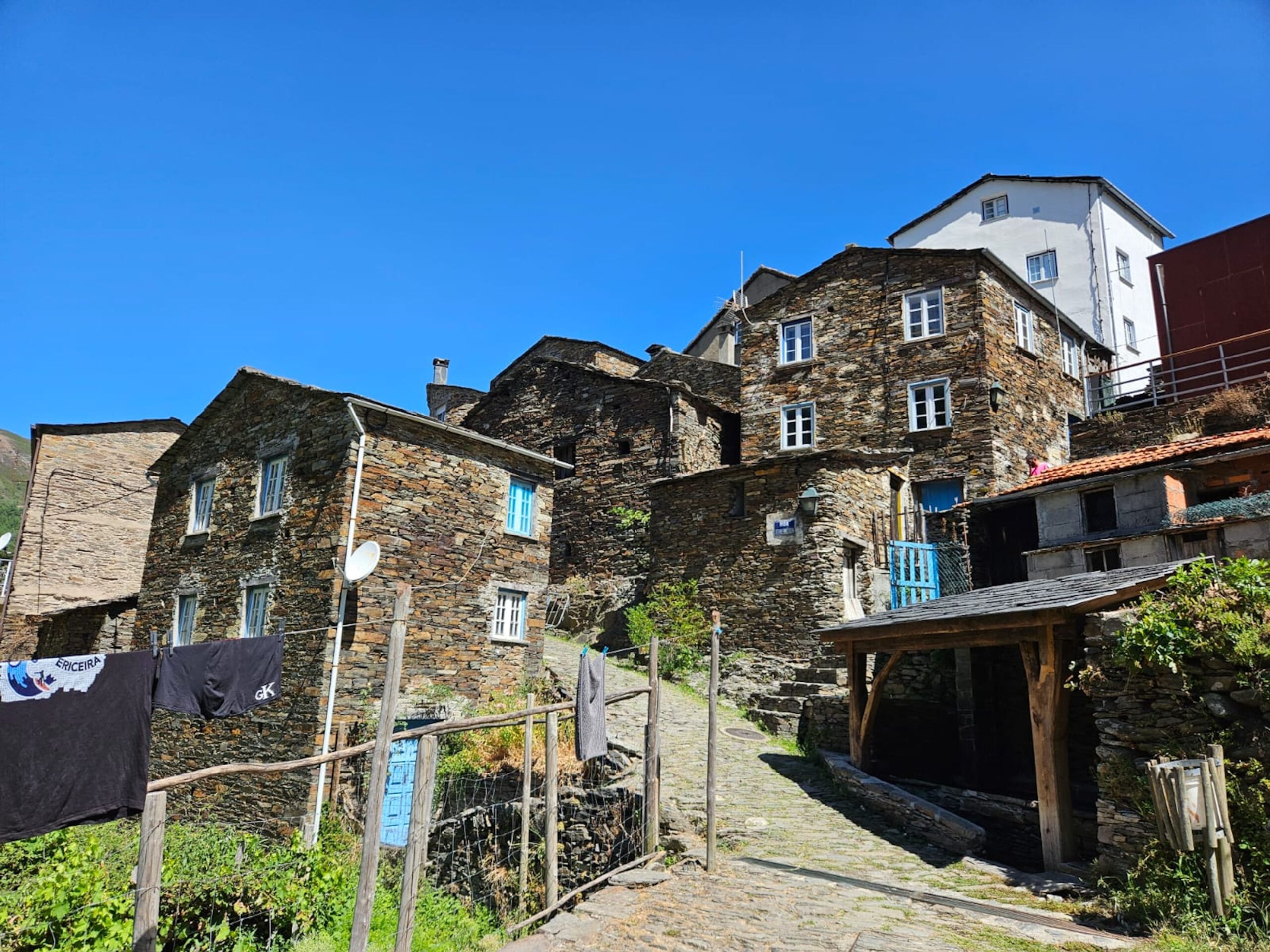 Fotografia em plano médio de casas tradicionais da Aldeia Histórica do Piódão em Portugal, construídas com pedras escuras de xisto e telhados de lousa, sob um céu azul limpo, ilustrando a crônica de Laura Moura Nunes.