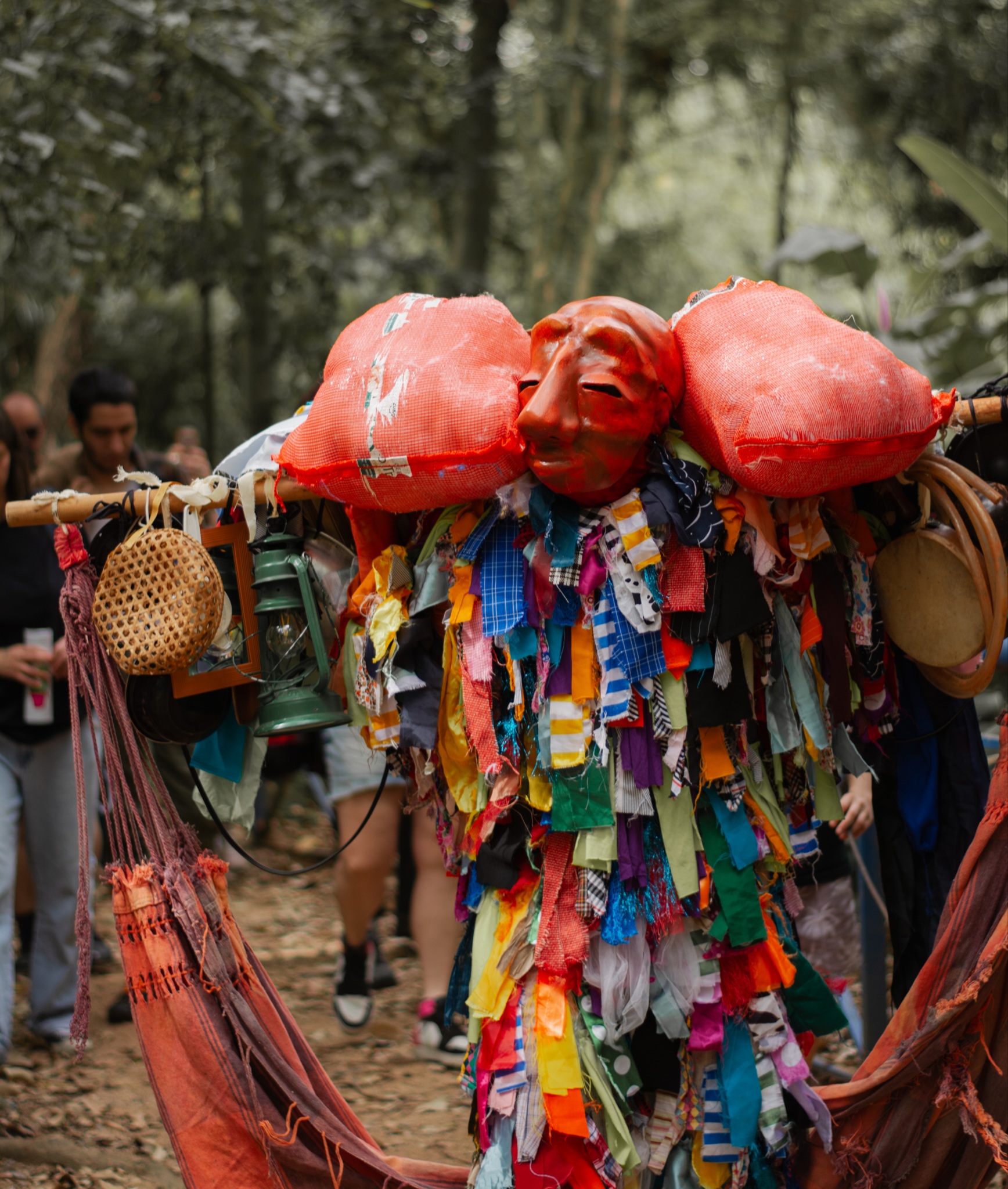 Artista performático em meio à natureza veste um traje folclórico feito com tiras de tecidos multicoloridos. Na altura dos ombros, uma estrutura de madeira carrega uma grande máscara vermelha de feições humanas, ladeada por dois sacos laranjas, além de objetos pendurados como um lampião verde, um cesto de palha e um pandeiro. Ao fundo, o público caminha por uma trilha cercada pelas árvores do Parque Natural da Biquinha.
