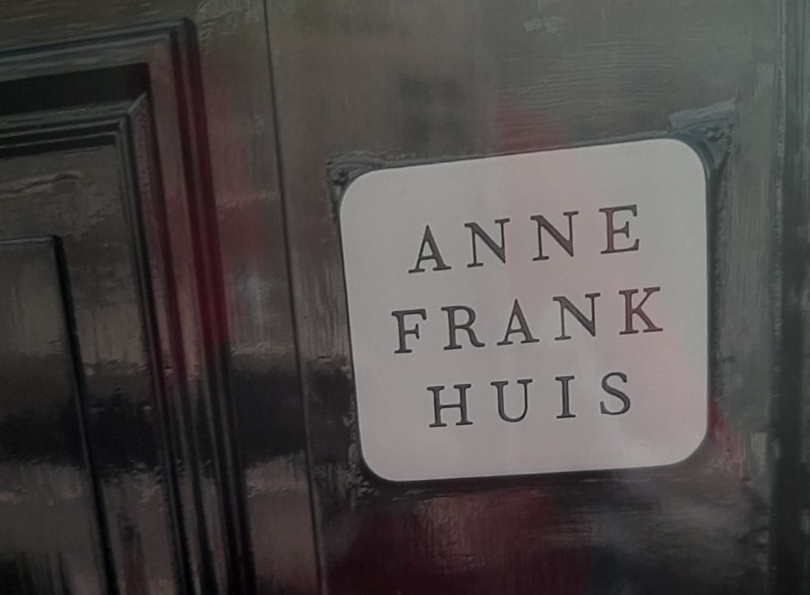 Uma placa branca com cantos arredondados e letras maiúsculas pretas onde se lê "ANNE FRANK HUIS", afixada numa superfície escura e brilhante, possivelmente uma porta.