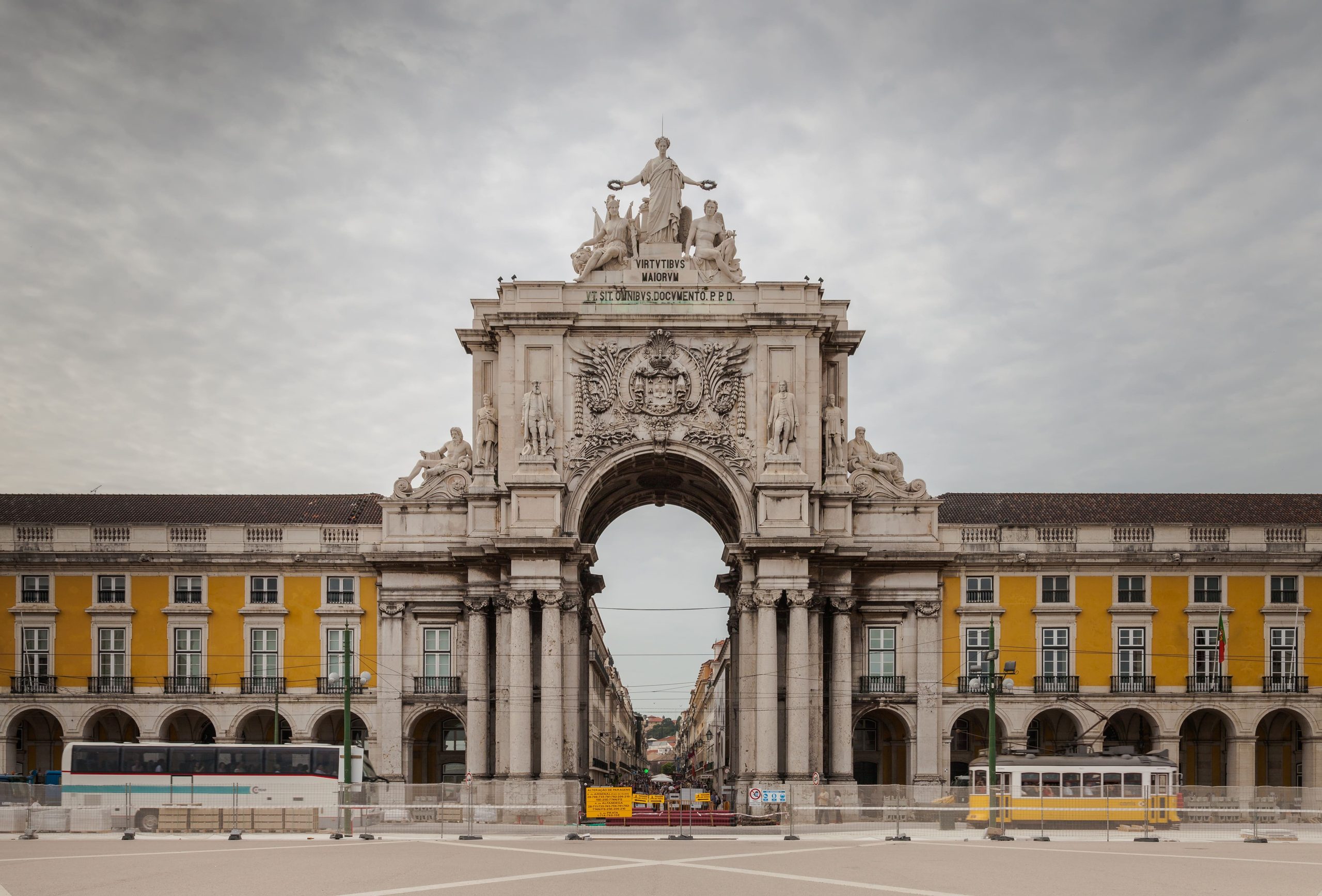 Fotografia do Arco da Rua Augusta, na Praça do Comércio, em Lisboa. O imponente arco de pedra branca liga os edifícios amarelos neoclássicos, com estátuas no topo e um elétrico amarelo a passar em baixo.