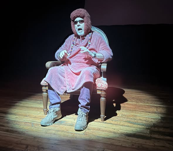 Uma pessoa com barba grisalha e óculos escuros sentada numa cadeira num palco de teatro sob a luz de um refletor. Veste um ropão ou túnica cor-de-rosa/aveludada sobre calças, um chapéu ou gorro acolchoado/peludo com abas para as orelhas e botas claras. Tem também colares coloridos e está com as mãos levantadas à altura do peito, expressando-se com a boca aberta.