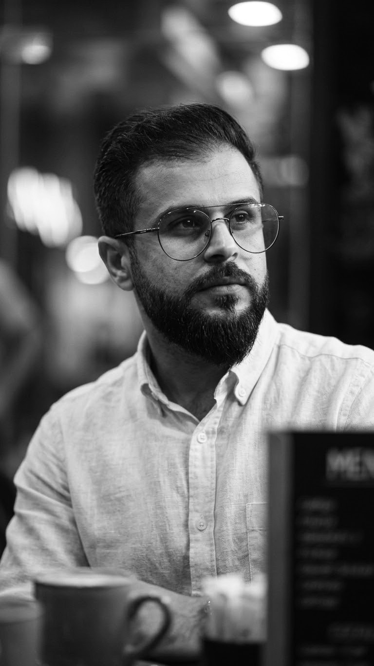 Maliki Al-Batli - Um homem jovem de óculos e barba e cabelos pretos, vestido com uma camisa branca